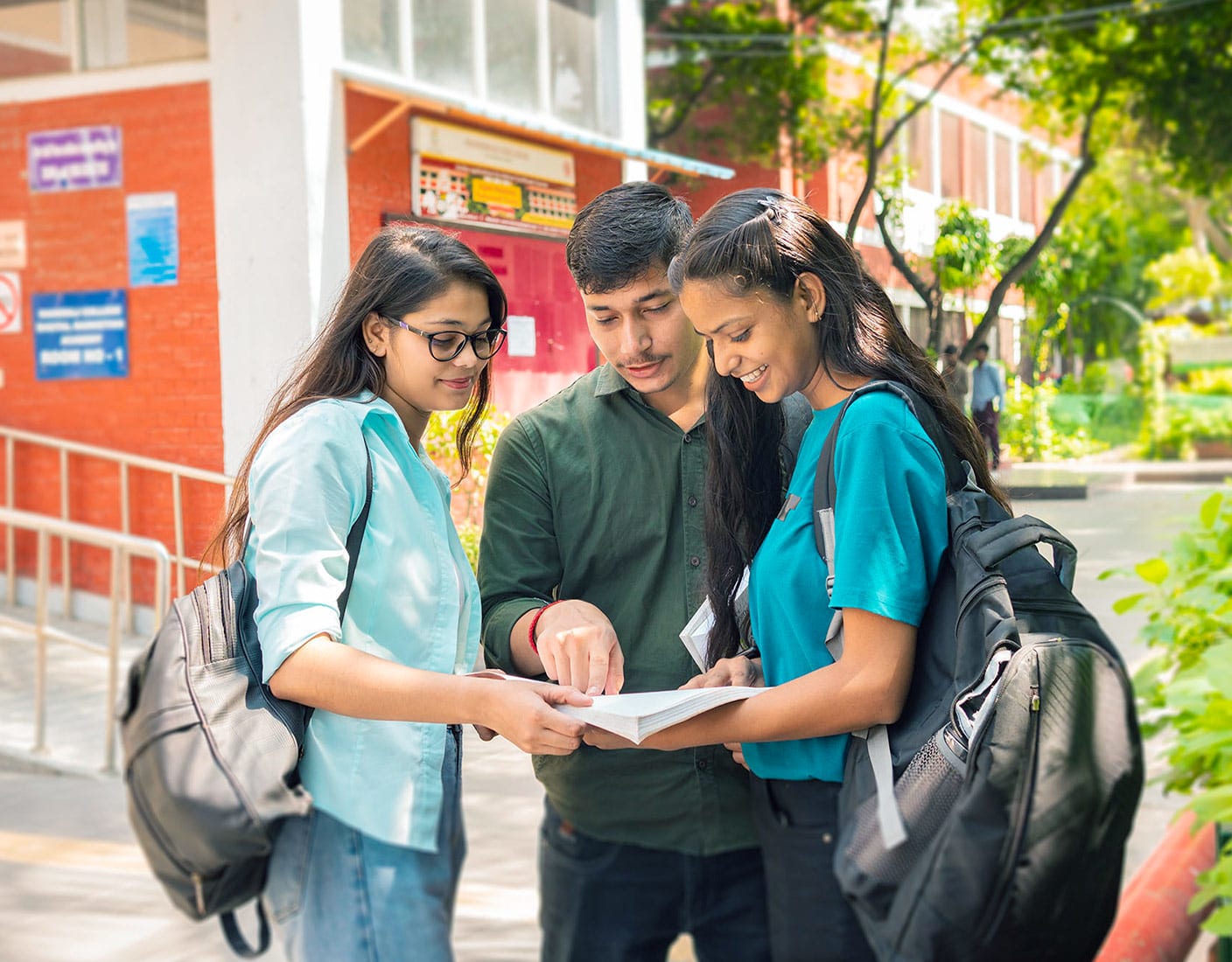 Three students with backpacks gathered outside classroom studying a textbook.
